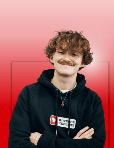 Junger Mann mit lockigem Haar und Schnurrbart, trägt ein schwarzes Hoodie mit Logo. Er lächelt vor einem rosa-roten Hintergru