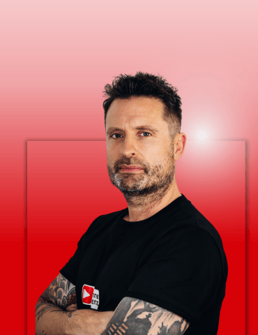 Mann mit kurzem, gestuften Haar und Bart, trägt schwarzes T-Shirt, steht mit verschränkten Armen vor einem rotgradienten Hint