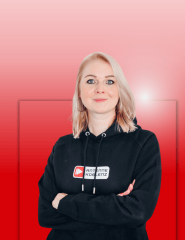 Frau mit blonden Haaren in schwarzem Hoodie steht mit verschränkten Armen vor einem einfarbigen, roten Hintergrund.