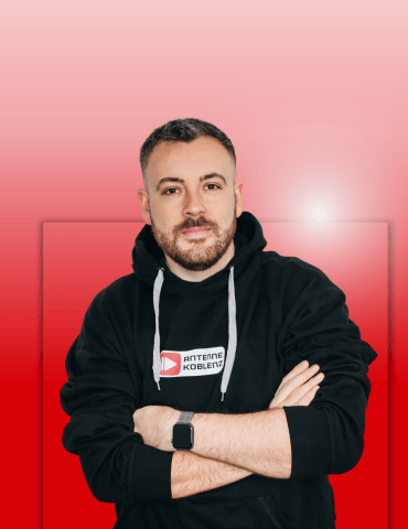 Mann mit kurzen Haaren und Bart in schwarzem Hoodie, stehend mit verschränkten Armen vor einem rot-rosa Farbverlauf.