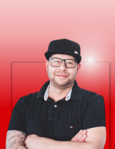 Mann mit Brille und schwarzem Hut, im schwarzen Polohemd, lächelt vor einem rot-verlaufenden Hintergrund.