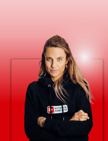 Frau mit langen, blonden Haaren trägt einen schwarzen Hoodie mit Logo. Sie steht vor einem roten Hintergrund.