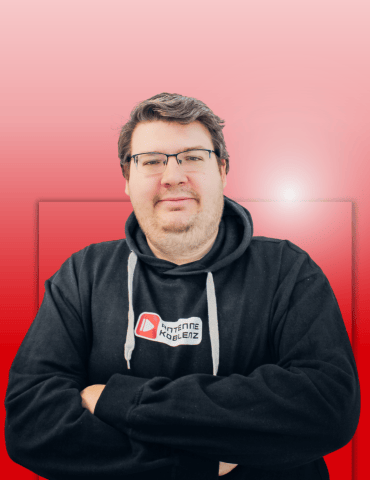 Ein Mann mit Brille trägt einen schwarzen Hoodie und steht mit verschränkten Armen vor einem rosa Hintergrund.