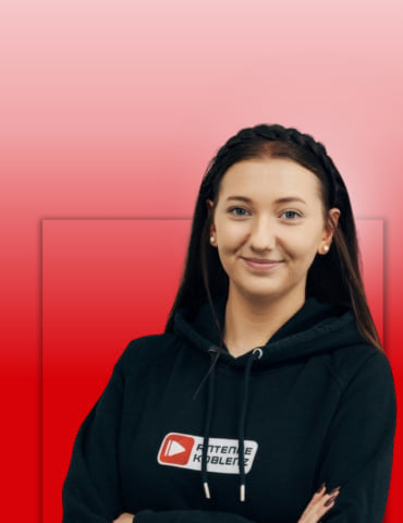 Eine lächelnde Frau mit mittellangen, braunen Haaren trägt einen schwarzen Hoodie mit Logo. Der Hintergrund ist rot.